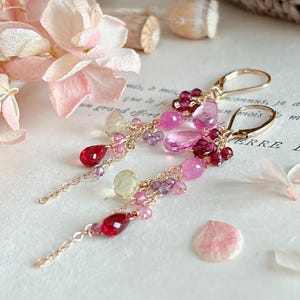 May include: Gold-tone dangle earrings with a cluster of pink, red, and purple gemstones. The earrings feature a lever-back closure and delicate chain accents. The gemstones include teardrop and round shapes, with a floral arrangement in the background.