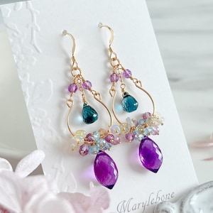 以下が含まれることがあります： ゴールドトーンのイヤリング。下部に涙型の紫色の宝石が付いています。イヤリングには、紫、青、白の色合いの小さなビーズのクラスターがあります。イヤリングは白いカードに表示されています。