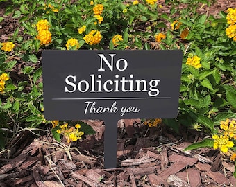 May include: Black sign with white text that reads "No Soliciting Thank you" placed in a bed of brown mulch and yellow flowers.