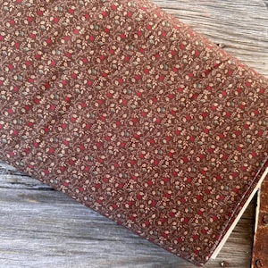 Puede incluir: Un rollo de tela con un estampado floral repetido. La tela es de color marrón oscuro, cubierta con pequeños diseños florales y de hojas rojas, blancas y verdes. La tela descansa sobre una superficie de madera envejecida.