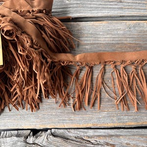 May include: Close-up of brown suede fringe trim and a matching piece of fabric. The fringe is long and flowing, with a raw edge. A small tag with a sewing machine illustration hangs from the fabric.
