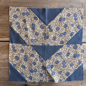 May include: A square fabric block with a floral pattern in blue, green, and cream, framed by solid blue-gray triangles. The block is likely part of a quilt or other textile project, showcasing a geometric design.