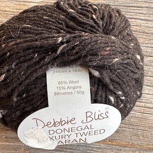 May include: A ball of dark brown Donegal Luxury Tweed Aran yarn by Debbie Bliss. The yarn is 85% wool and 15% angora, with white and brown flecks. The label indicates 88 metres per 50g. Suitable for knitting and crochet projects.