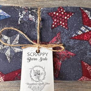 Pode incluir: Um tecido dobrado com um padrão de estrelas em vermelho, branco e azul, amarrado com barbante e uma etiqueta que diz "SCRAPPY Barn Sale". O tecido tem um fundo azul escuro com vários desenhos de estrelas.