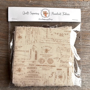 May include: A package of quilt squares with a vintage French script design. The fabric is beige with brown text and illustrations, including bees and the word "Paris". The package is labeled "Quilt Squares Handcut Fabric".