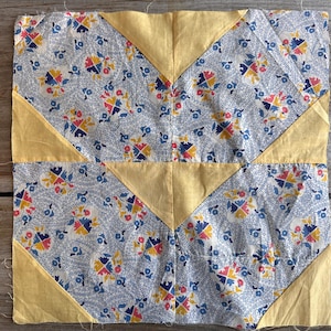 May include: A square quilt block with a floral pattern in blue, red, and yellow on a white background. The block features yellow triangles at the corners and along the edges, creating a geometric design. The fabric appears to be cotton.