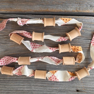 Może przedstawiać: Ręcznie robiona girlanda z drewnianych szpulek i pasków materiału. Materiał ma mieszankę wzorów, w tym czerwone kwiaty i pomarańczowe kropki. Girlanda jest ułożona na zwietrzałej drewnianej powierzchni.