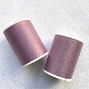 May include: Two spools of lavender-colored thread. The thread is wound around a cylindrical core with white edges. The spools are positioned at a slight angle against a white, patterned background. The thread has a subtle sheen.