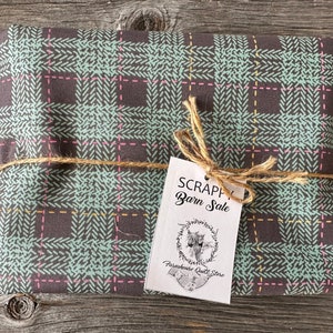 Puede incluir: Un paquete envuelto en tela de cuadros marrones y verdes con una etiqueta que dice "Scrappy Barn Sale" y "Farmhouse Quilt Store".