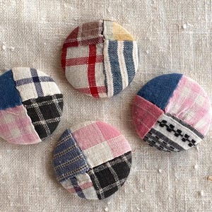 May include: Four handmade, circular fabric buttons with a patchwork design. Each button is divided into four sections of different patterned fabrics, including plaid, stripes, and solid colors in shades of red, blue, pink, black, and white.