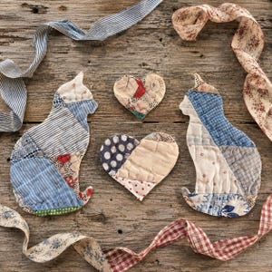May include: Two quilted fabric cat silhouettes and two heart-shaped fabric ornaments featuring a patchwork of blue, white, and red patterned textiles. The cat and heart shapes are arranged with flowing fabric ribbons in blue stripes and floral prints.
