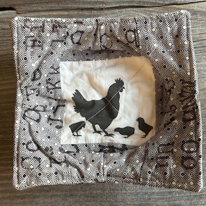 Puede incluir: Funda cuadrada de tela para cuenco con un estampado geométrico en blanco y negro. El centro presenta un cuadrado blanco con una gallina negra y tres pollitos. La funda está diseñada para sujetar un cuenco de comida caliente.