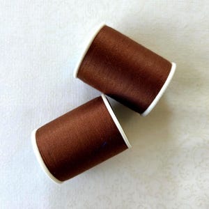 May include: Two spools of brown thread with white edges are displayed against a white background. The thread is tightly wound on the spools, and the color is a rich, dark brown. The spools are positioned at a slight angle to each other.