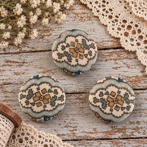 May include: Three circular fabric-covered buttons featuring a repeating pattern in shades of muted blue, cream, and mustard yellow. Each button has a soft, rounded surface with a textile texture, displaying a symmetrical floral-inspired design.