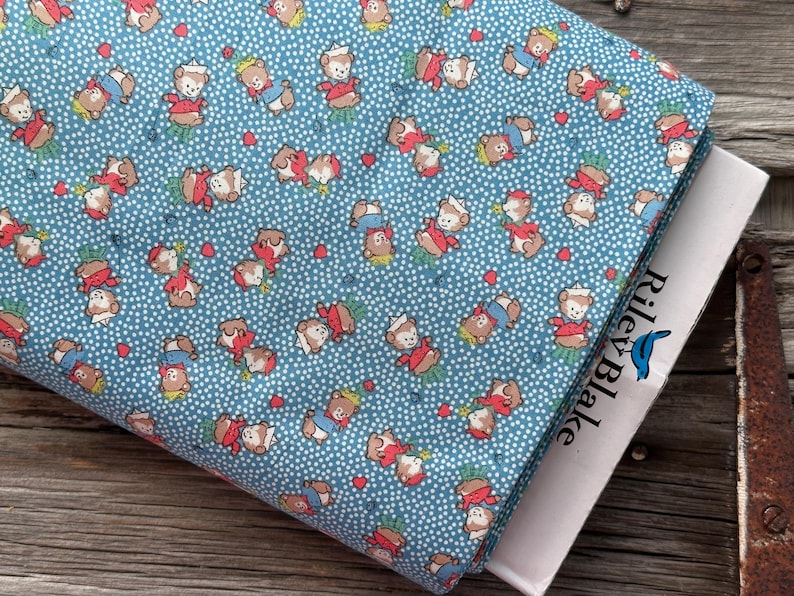 May include: A folded fabric featuring a repeating pattern of cartoon teddy bears in red, white, and blue outfits on a light blue background with white polka dots. The fabric is partially covering a book with the text "Riley Blake".