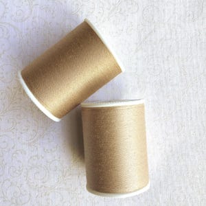 May include: Two spools of gold-colored thread are displayed against a white background with a subtle floral pattern. The thread spools have white rims and are positioned at an angle, showcasing the metallic sheen of the thread.