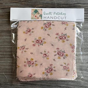 Puede incluir: Un paquete de parches de edredón cortados a mano. La tela tiene un fondo rosa claro con un estampado floral de rosas rosas y amarillas. El texto "Quilt Patches HANDCUT" está impreso en el embalaje.