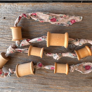 May include: A garland of wooden spools connected by fabric strips. The fabric is a light beige with a floral pattern of red and pink roses. The spools are made of light-colored wood.