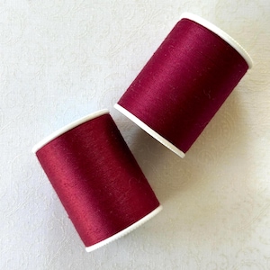 May include: Two spools of burgundy thread with white rims are displayed against a white, textured background. The thread is tightly wound, and the spools are positioned at a slight angle, showcasing the rich color and texture of the thread.