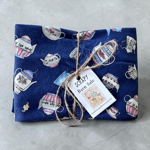 Puede incluir: Un paquete de tela azul marino doblado, con un patrón repetido de teteras blancas, rosas y azules. Una etiqueta dice "SCRAPY Barn Sale" con una pequeña ilustración, atada con hilo.