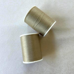 May include: Two spools of metallic gold thread with white rims are displayed against a light background. The thread is tightly wound on the spools, and the color has a subtle sheen. The spools are positioned at an angle, creating a visually appealing arrangement.