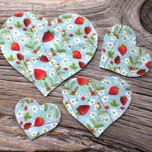 May include: Five heart-shaped fabric patches with a light blue background. The patches feature a strawberry and floral pattern with red stitching. The strawberries are red, and the flowers are white.