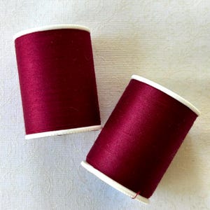 May include: Two spools of burgundy thread with white rims. The thread is tightly wound on cylindrical spools, showcasing a rich, deep red color. The spools are positioned on a white, textured surface, suggesting a sewing or crafting theme.