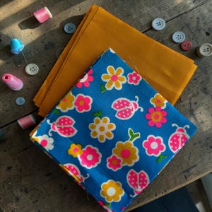 Può includere: Un assortimento di articoli per cucire su una superficie di legno. Include rocchetti di filo rosa e blu, vari bottoni e quadrati di tessuto. Uno è blu con un motivo floreale e coccinelle, l'altro è giallo senape.