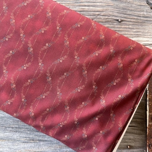 Puede incluir: Un trozo de tela doblado en un tono rojo intenso, adornado con un patrón repetitivo de delicadas enredaderas florales. Las enredaderas están acentuadas con pequeños elementos florales azules y dorados, creando un diseño sutil y elegante. La tela es para coser o manualidades.