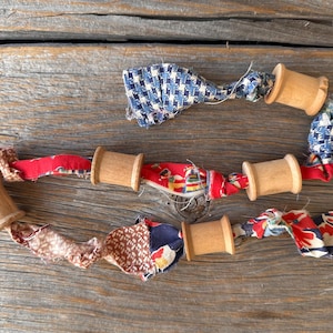 Może przedstawiać: Ręcznie robiona girlanda z drewnianych szpulek połączonych kolorowymi paskami materiału. Materiał prezentuje wzory w kolorach czerwonym, niebieskim i brązowym, tworząc rustykalną i uroczą estetykę. Girlanda leży na zwietrzałej drewnianej powierzchni.