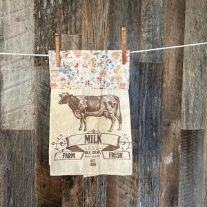 May include: A beige kitchen towel with a floral top and a cow illustration. The towel has the words "Milk, Pure, Cream, Fresh, Farm" printed on it. The towel is hanging on a clothesline with wooden clothespins.