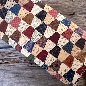 Pode incluir: Um edredom dobrado com um design de patchwork. O edredom apresenta uma variedade de tecidos em um padrão de losangos, com cores incluindo vermelho, azul, marrom e creme. O edredom está sobre uma superfície de madeira.