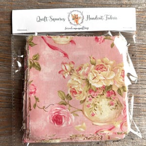 May include: A package of quilt squares featuring a pink floral pattern with roses and a vase. The package is labeled "Quilt Squares Handcut Fabric" with a logo. The fabric has a vintage, romantic aesthetic.
