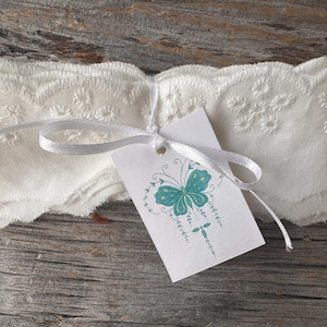 May include: A bundle of white, embroidered fabric with a scalloped edge, tied with a white ribbon. A tag with a teal butterfly design is attached. The fabric is folded and appears to be made of a soft material.