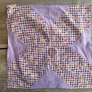May include: A square patchwork quilt block with a purple and white gingham pattern, accented with floral details in orange, red, and yellow. The block features four lavender triangles at the corners and center.