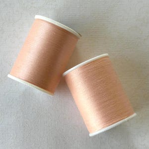 May include: Two spools of peach-colored thread with white rims. The thread is tightly wound on cardboard spools. The spools are positioned at a slight angle against a white, textured background. Ideal for sewing and crafting projects.