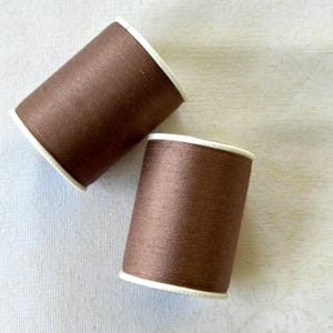 May include: Two spools of brown thread with white rims are angled on a white patterned surface. The thread appears to be a medium brown color, and the spools are cylindrical in shape. The image is well-lit, highlighting the texture of the thread.