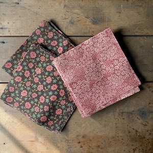 Puede incluir: Tres servilletas de tela plegadas con estampados florales. Dos servilletas presentan un fondo gris oscuro con flores rosas y hojas verdes. La tercera tiene un fondo rosa con un estampado floral blanco. Las servilletas están sobre una superficie de madera.