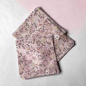 Può includere: Una pila di tovaglioli di stoffa rosa a fantasia floreale. I tovaglioli presentano un delicato motivo di piccoli fiori e foglie bianche, gialle e marroni. Il tessuto è piegato a forma quadrata, con diversi tovaglioli impilati.