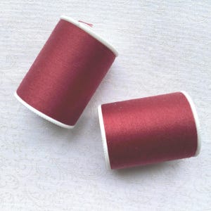 May include: Two spools of burgundy thread with white rims are displayed against a white, patterned background. The thread appears to be a rich, solid color, and the spools are cylindrical in shape. The image is well-lit, highlighting the texture of the thread.
