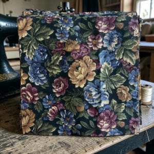Può includere: Una pila di quadrati di tessuto floreale con uno sfondo scuro e un motivo di fiori blu, oro e bordeaux e foglie verdi. Il tessuto è probabilmente cotone o un materiale simile, adatto per progetti di quilting o artigianato.