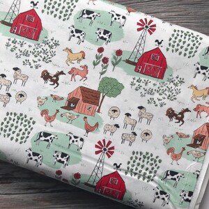 May include: White fabric with a repeating farm scene pattern. The design includes red barns, windmills, cows, horses, sheep, chickens, and flowers. The fabric has a light green and red color scheme.