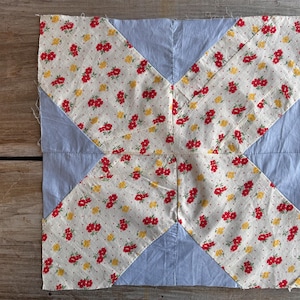May include: A square quilt block with a light blue background and four white sections with a red floral pattern. The quilt block is made of fabric and has a patchwork design.