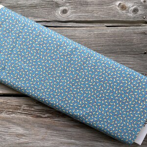 May include: A roll of blue fabric with a repeating pattern of small, white and yellow shapes. The fabric is labeled "Riley Blake Designs" on the end. The fabric is resting on a wooden surface.