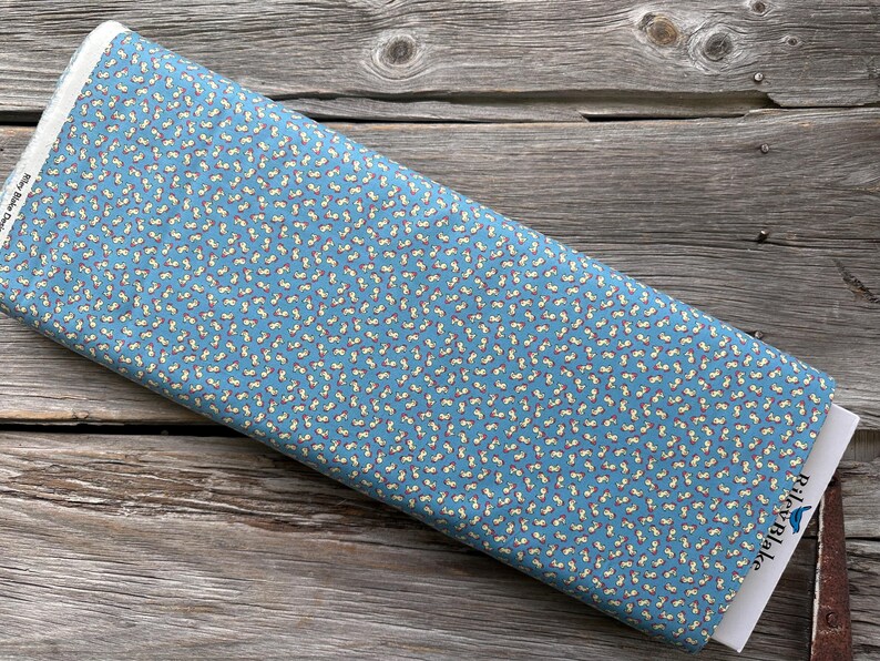 May include: A roll of blue fabric with a repeating pattern of small, white and yellow shapes. The fabric is labeled "Riley Blake Designs" on the end. The fabric is resting on a wooden surface.