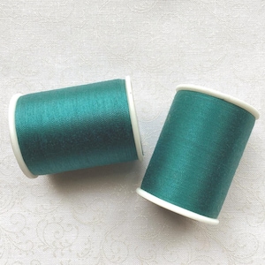 May include: Two spools of teal thread with white rims are displayed against a white patterned background. The thread is tightly wound, and the spools are cylindrical. The color is a deep, rich teal, and the thread appears to have a slight sheen.