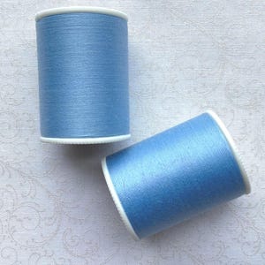 May include: Two spools of light blue thread with white rims. The spools are cylindrical and appear to be full. The thread has a subtle sheen. The background is a white fabric with a subtle pattern.