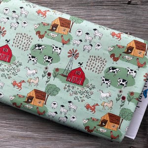 May include: A roll of fabric with a farm-themed pattern. The design features red barns, windmills, cows, sheep, horses, and chickens on a light green background. The fabric is folded and resting on a wooden surface.