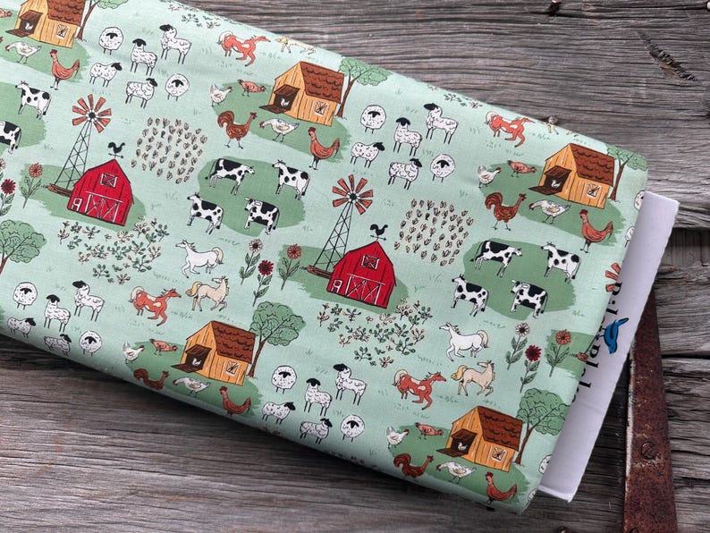 May include: A roll of fabric with a farm-themed pattern. The design features red barns, windmills, cows, sheep, horses, and chickens on a light green background. The fabric is folded and resting on a wooden surface.