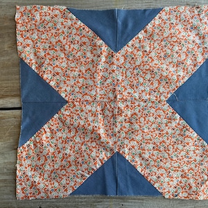 May include: A square fabric block with a patchwork design. The center features a floral pattern in orange, red, and white. Blue fabric forms an X shape across the block. The block is likely part of a quilt or other textile project.